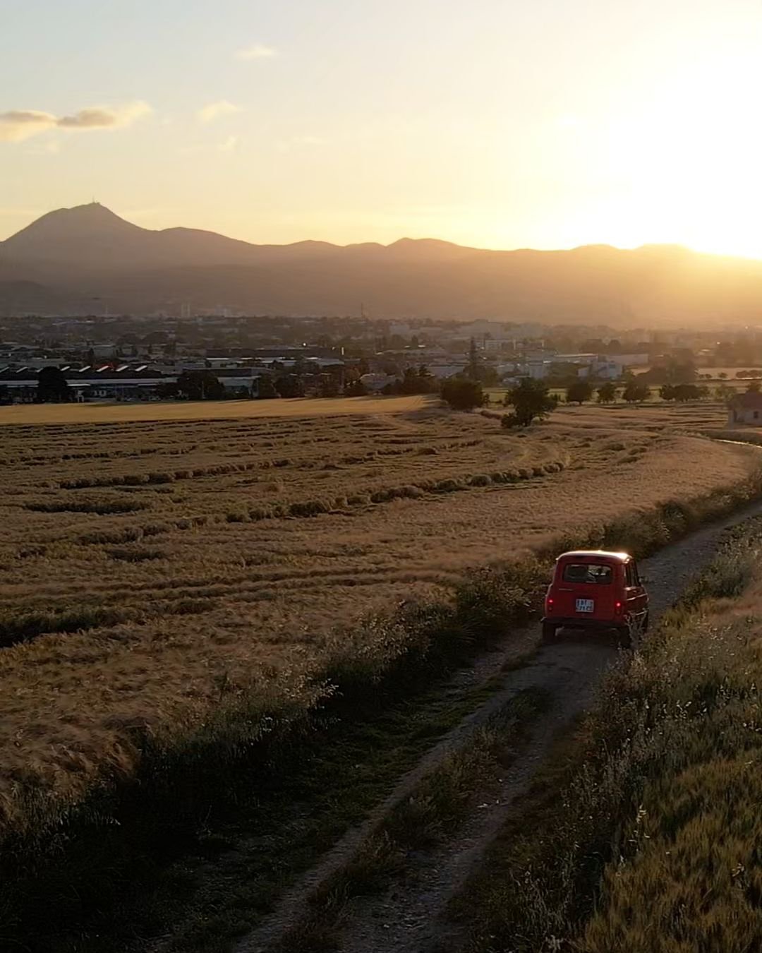 Vue de drone au coucher du soleil : une 4L rouge sur un chemin entre les champs, la chaîne des Puys à l’horizon
